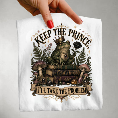 A hand with red nail polish holds up a white t-shirt featuring a whimsical graphic of a crowned frog sitting on books, surrounded by mushrooms, ferns, and a rose, with the text 'KEEP THE PRINCE, I'LL TAKE THE PROBLEM'.