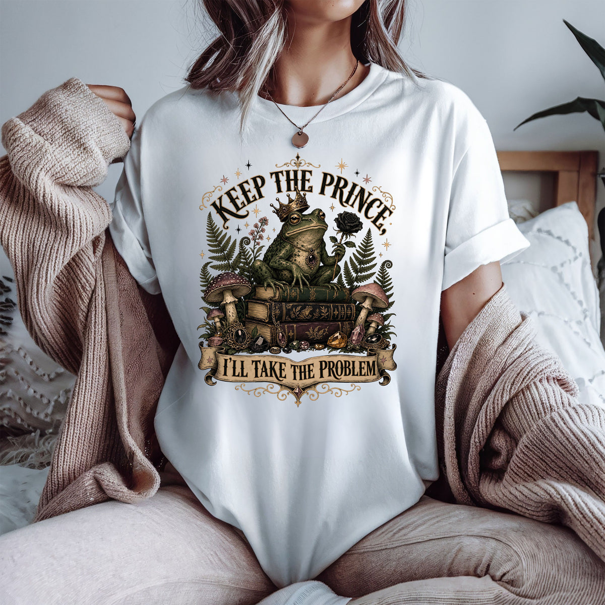 A woman wearing a white graphic t-shirt with a vintage-style frog illustration and the text 'KEEP THE PRINCE' and 'I'LL TAKE THE PROBLEM'. She is sitting on a bed with a cozy beige knit sweater draped off her shoulders.