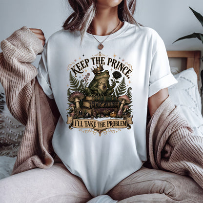 A woman wearing a white graphic t-shirt with a vintage-style frog illustration and the text 'KEEP THE PRINCE' and 'I'LL TAKE THE PROBLEM'. She is sitting on a bed with a cozy beige knit sweater draped off her shoulders.