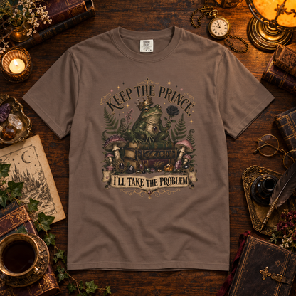 A brown graphic t-shirt displayed in a styled flat-lay on a dark wooden table surrounded by vintage props like a cup of tea, a pocket watch, books, and crystals. The t-shirt features a detailed fantasy graphic of a green frog sitting on a chest with the text 'KEEP THE PRINCE' above and 'I'LL TAKE THE PROBLEM' below.