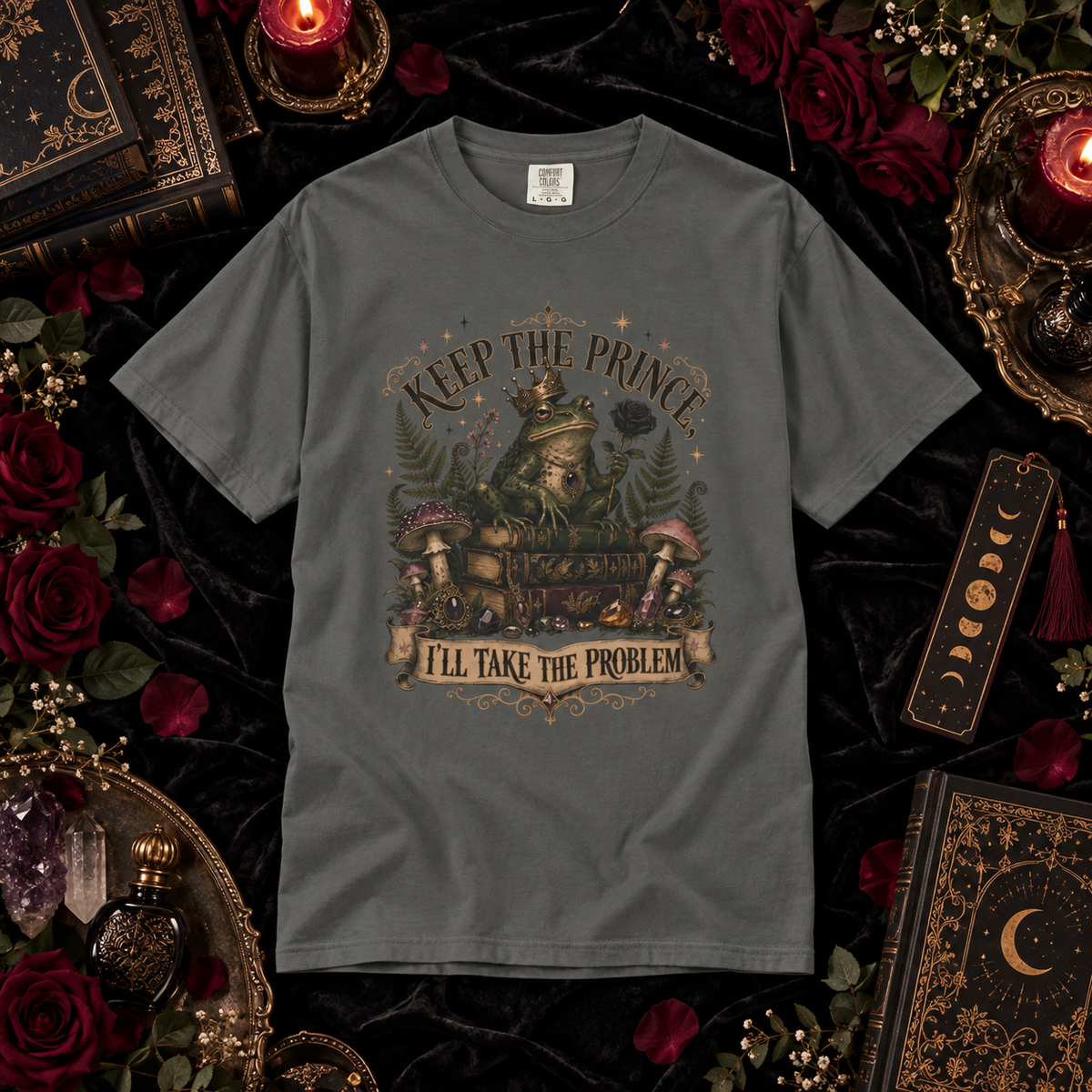 A dark gray graphic t-shirt laid flat on a black velvet background, surrounded by decorative props including red roses, candles, old books, crystals, and a tassel bookmark. The shirt features a detailed fantasy illustration of a princess in a carriage with mushrooms and the text 'KEEP THE PRINCE I'LL TAKE THE PROBLEM'.
