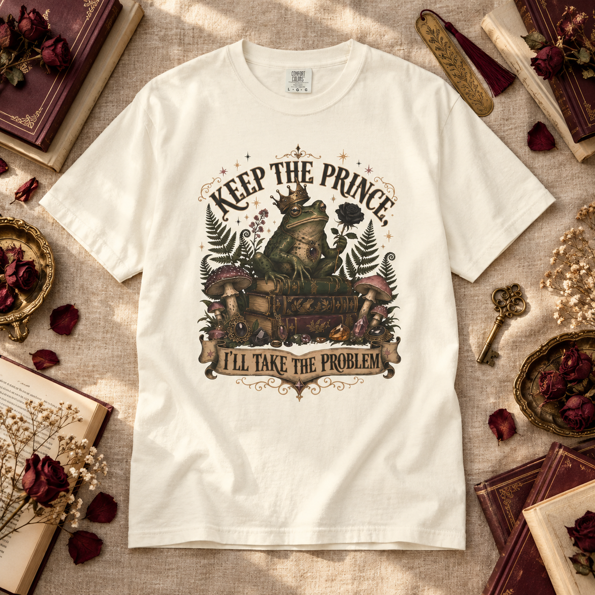 A white graphic t-shirt laid flat on a textured beige fabric background, surrounded by vintage books, dried red roses, and a key. The t-shirt features a detailed fantasy illustration of a frog wearing a crown and holding a chest, with the text 'KEEP THE PRINCE' above and 'I'LL TAKE THE PROBLEM' below.