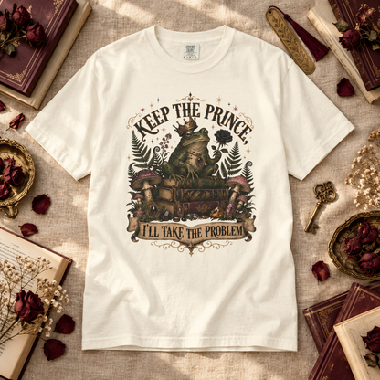 A white graphic t-shirt laid flat on a textured beige fabric background, surrounded by vintage books, dried red roses, and a key. The t-shirt features a detailed fantasy illustration of a frog wearing a crown and holding a chest, with the text 'KEEP THE PRINCE' above and 'I'LL TAKE THE PROBLEM' below.