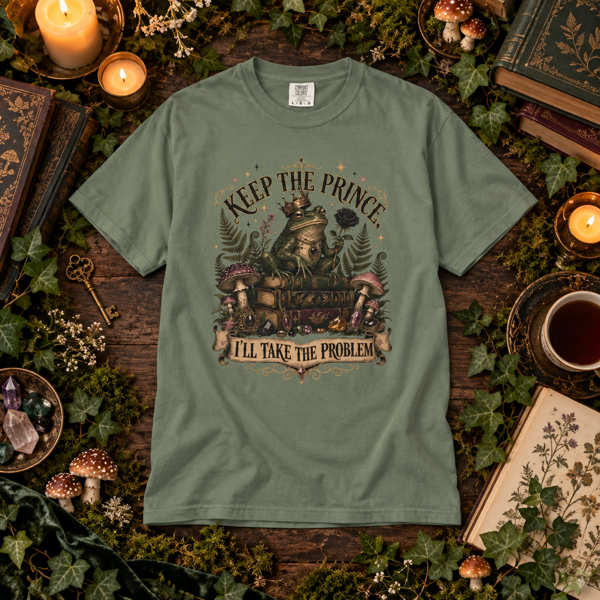 An olive green graphic t-shirt laid flat on a rustic wooden surface surrounded by moss, candles, mushrooms, books, and a cup of tea. The t-shirt features a detailed graphic of a frog wearing a crown and a train, with the text 'KEEP THE PRINCE, I'LL TAKE THE PROBLEM'.