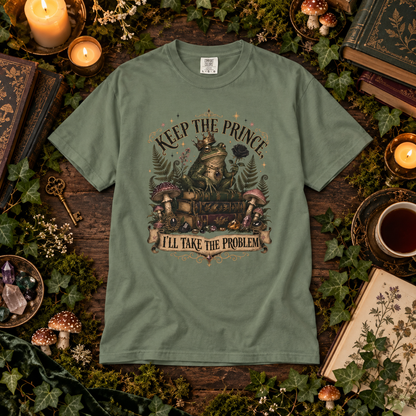 An olive green graphic t-shirt laid flat on a rustic wooden surface surrounded by moss, candles, mushrooms, books, and a cup of tea. The t-shirt features a detailed graphic of a frog wearing a crown and a train, with the text 'KEEP THE PRINCE, I'LL TAKE THE PROBLEM'.