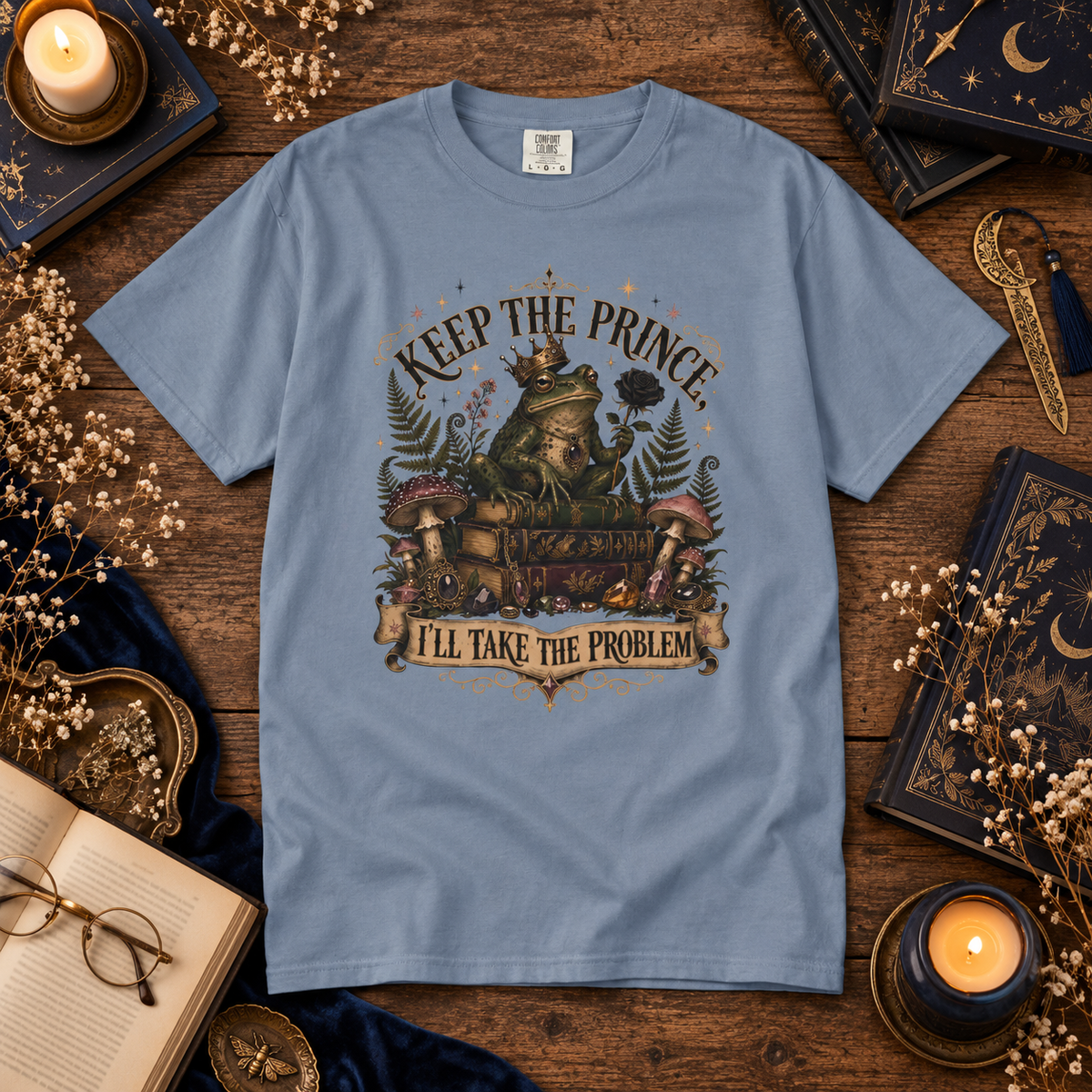 A light blue graphic t-shirt laid flat on a rustic wooden table surrounded by cozy props like candles, books, glasses, and dried flowers. The t-shirt features a detailed fantasy illustration of a frog wearing a crown and holding a book, with the text 'KEEP THE PRINCE' above and 'I'LL TAKE THE PROBLEM' below.