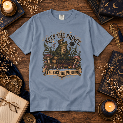 A light blue graphic t-shirt laid flat on a rustic wooden table surrounded by cozy props like candles, books, glasses, and dried flowers. The t-shirt features a detailed fantasy illustration of a frog wearing a crown and holding a book, with the text 'KEEP THE PRINCE' above and 'I'LL TAKE THE PROBLEM' below.