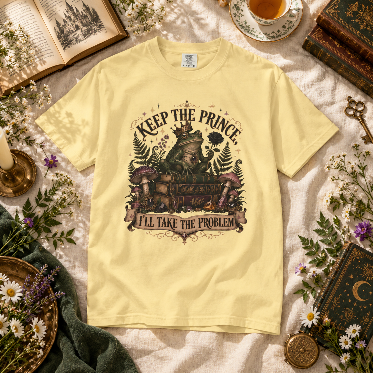 A pale yellow graphic T-shirt laid flat on a textured cream fabric, surrounded by styled props including an open book, teacup, key, flowers, and vintage books. The shirt features a large illustrated chest graphic with the text 'KEEP THE PRINCE, I'LL TAKE THE PROBLEM' and a fairy illustration.