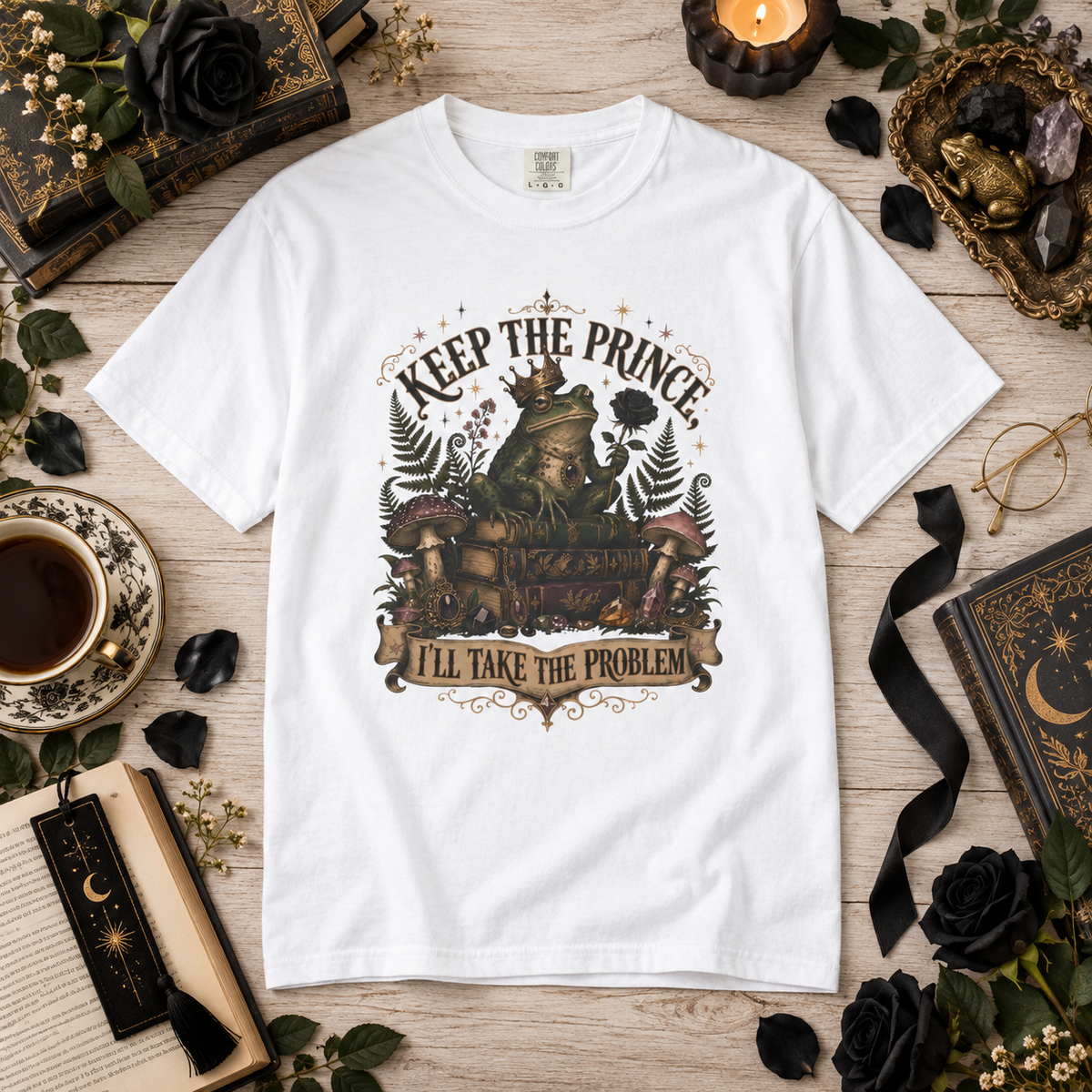 A white graphic t-shirt featuring a whimsical illustration of a princess in a crown sitting on a throne with the text 'KEEP THE PRINCE I'LL TAKE THE PROBLEM'. The t-shirt is displayed in a styled flat-lay on a light wood background surrounded by thematic props including black roses, a cup of coffee, a candle, and old books.