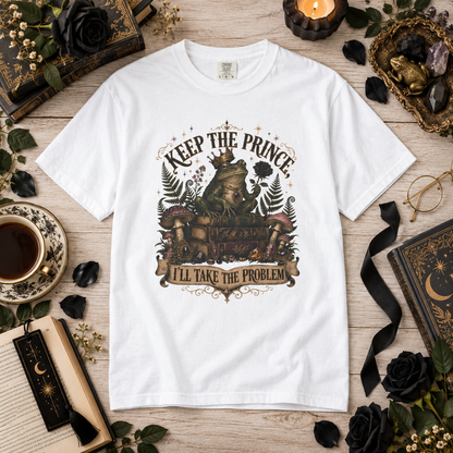 A white graphic t-shirt featuring a whimsical illustration of a princess in a crown sitting on a throne with the text 'KEEP THE PRINCE I'LL TAKE THE PROBLEM'. The t-shirt is displayed in a styled flat-lay on a light wood background surrounded by thematic props including black roses, a cup of coffee, a candle, and old books.