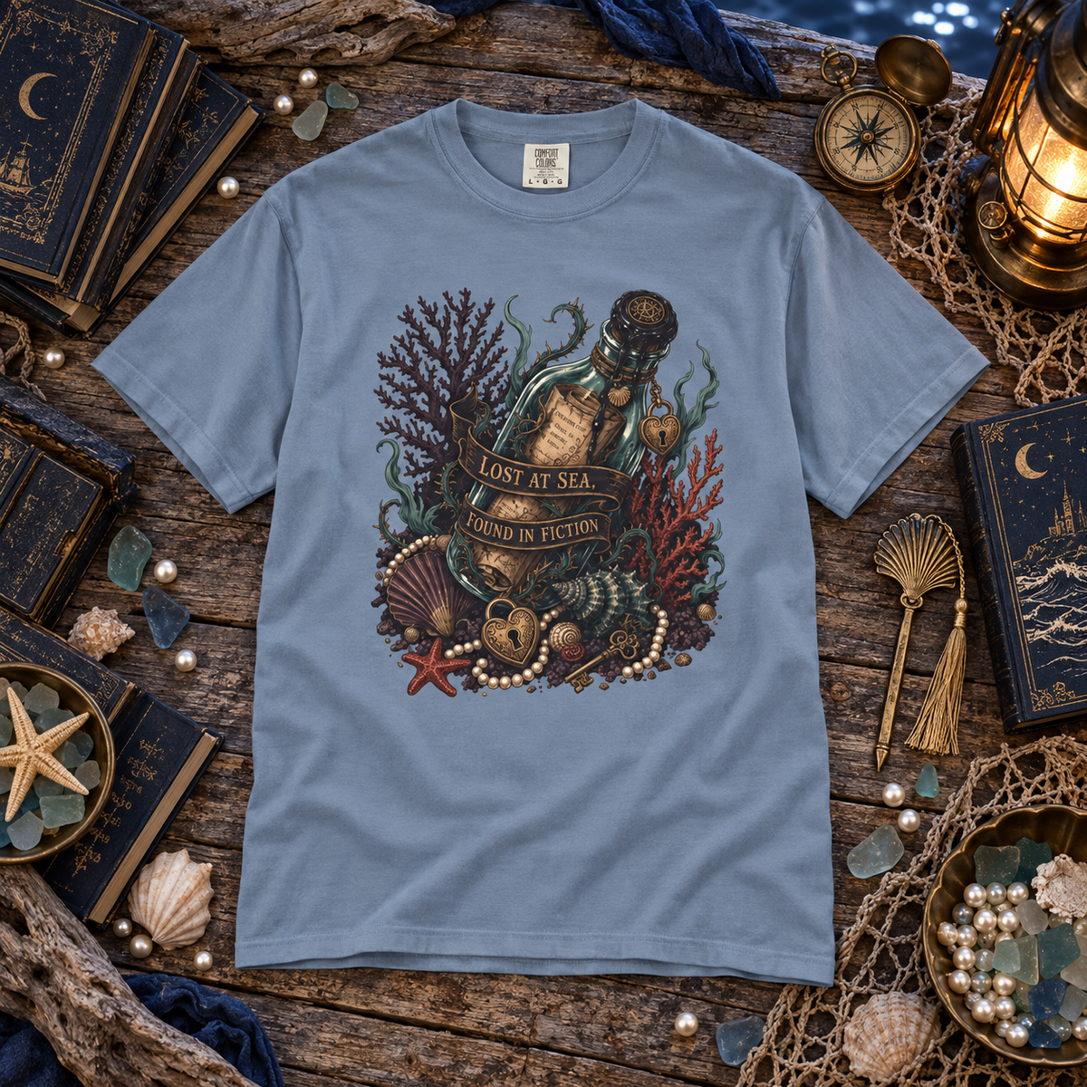A light blue crewneck t-shirt with a detailed graphic of an underwater scene featuring a bottle, coral, pearls, and a starfish. The shirt is displayed in a styled flat-lay on a rustic wooden surface surrounded by nautical props like a compass, lantern, shells, and books.