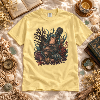 A pale yellow short-sleeve T-shirt laid flat on a textured cream fabric, surrounded by nautical props including shells, a compass, books, dried flowers, and a cup of tea. The shirt features a large central graphic of an underwater bottle sealed in sand with coral, shells, and a banner reading 'LOST AT SEA.