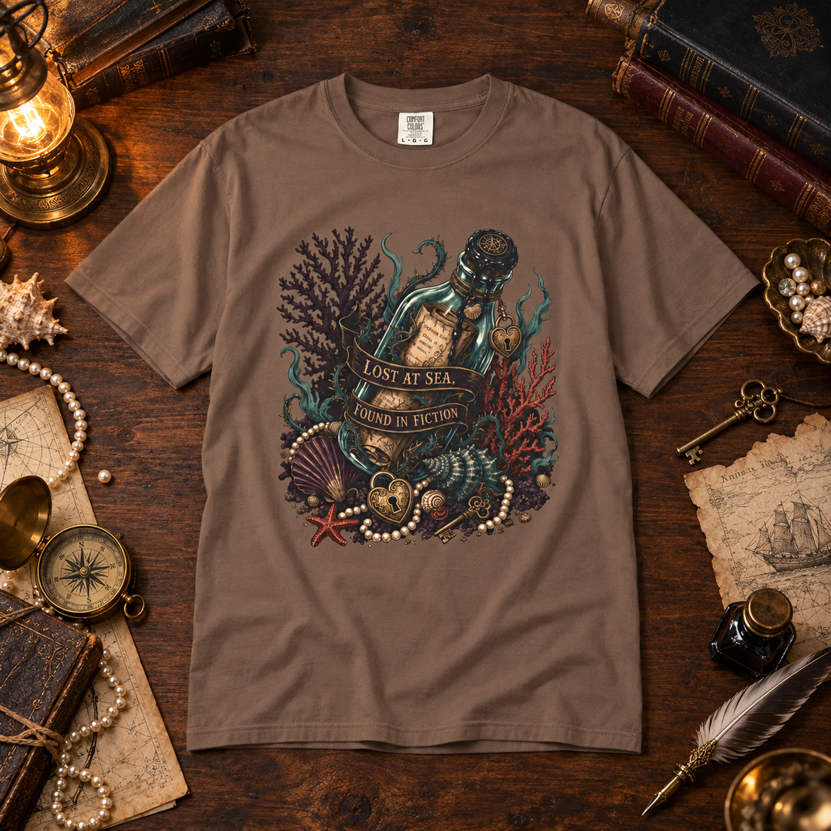 A brown short-sleeve graphic t-shirt laid flat on a dark wooden table surrounded by nautical and vintage props, including a lantern, books, pearls, a compass, and a feather quill. The t-shirt features a detailed graphic of a bottle sealed with a starfish, surrounded by sea corals, shells, and pearls, with the text 'LOST AT SEA FOUND IN FICTION'.