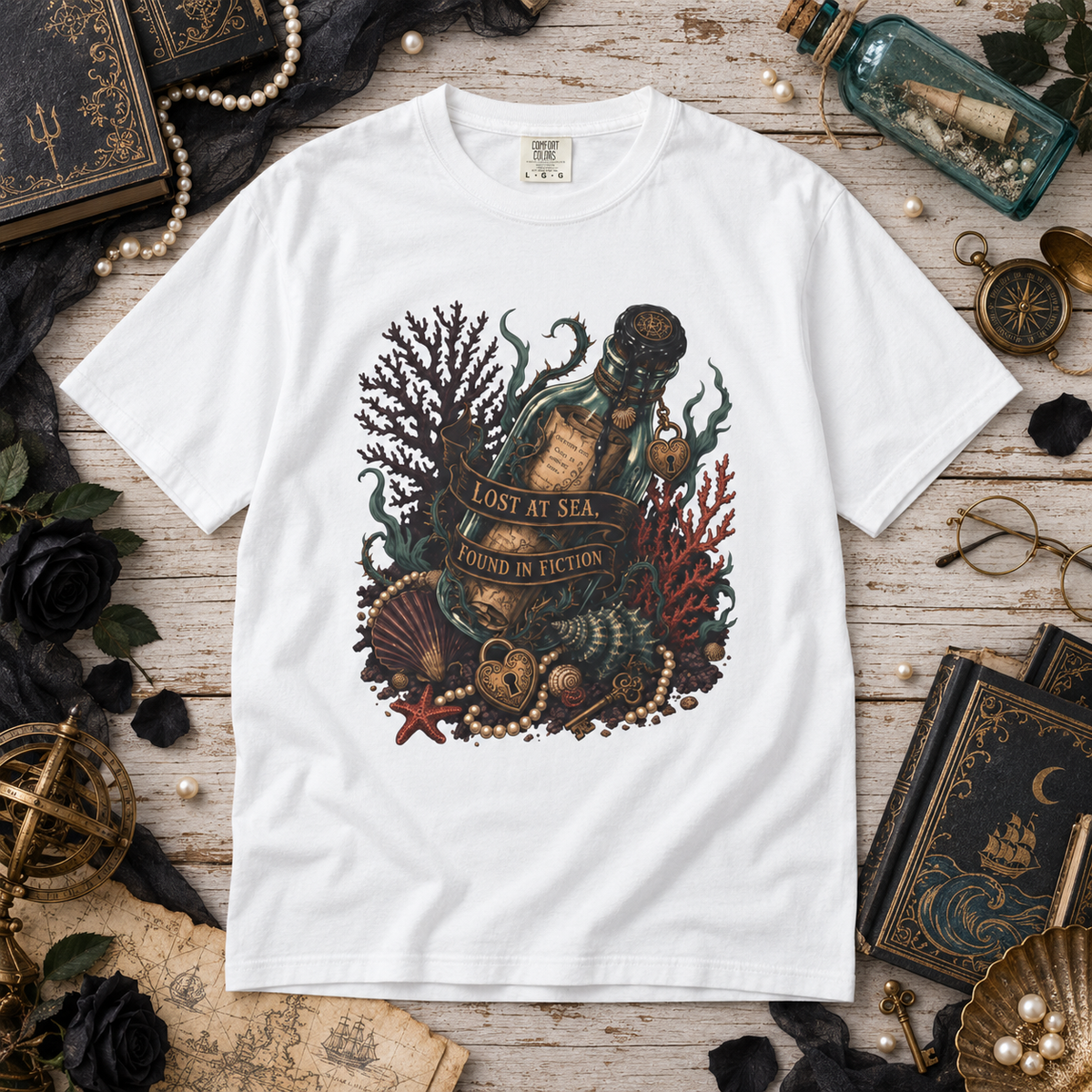 A white short-sleeve crewneck T-shirt laid flat on a rustic wooden surface, surrounded by nautical and vintage-themed props including books, pearls, a compass, glasses, a bottle, and a shell. The shirt features a large central graphic of an open bottle sealed with a letter, surrounded by sea urchins, coral, shells, and the text 'LOST AT SEA, FOUND IN FICTION'.