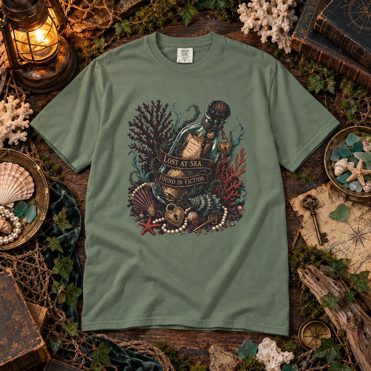 An olive green short-sleeve t-shirt with a detailed graphic of a glass bottle sealed with a cork, surrounded by sea urchins, pearls, coral, and a banner reading 'LOST AT SEA FOUND IN FICTION'. The shirt is displayed in a styled flat-lay setting with nautical props like a lantern, shells, and a compass on a rustic wooden background.