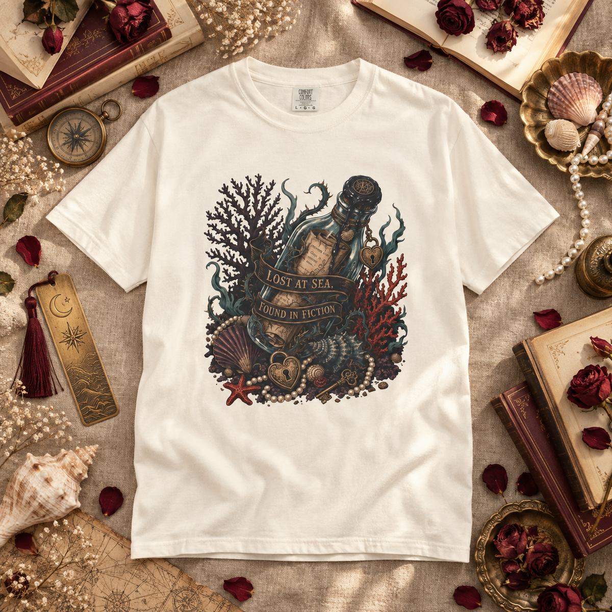 A white short-sleeve graphic T-shirt laid flat on a textured beige fabric surrounded by nautical and vintage props, including books, shells, pearls, dried flowers, and a compass. The shirt features a large central illustration of an underwater bottle scene with coral, starfish, shells, and a banner reading 'LOST AT SEA AND FOUND IN ECTION'.