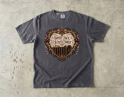 A dark gray heather t-shirt laid flat on a textured concrete surface, featuring a large heart-shaped graphic with the text 'SWEET FACE, SAVED SHELF' and a barbell design.