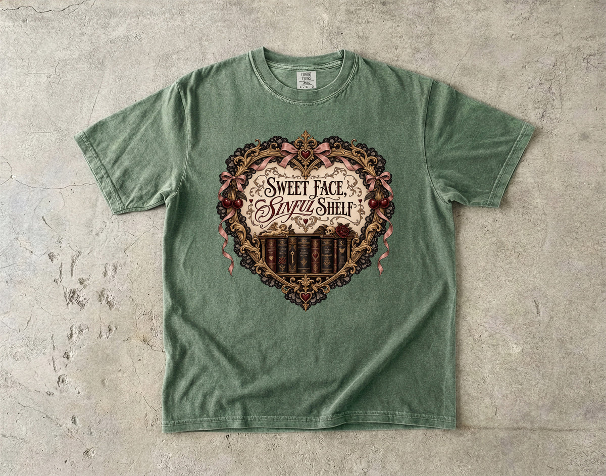 A sage green crewneck t-shirt laid flat on a textured concrete surface, featuring a large vintage-style heart graphic on the chest with the text 'Sweet Face, Sweet Shelf' and a row of book spines.