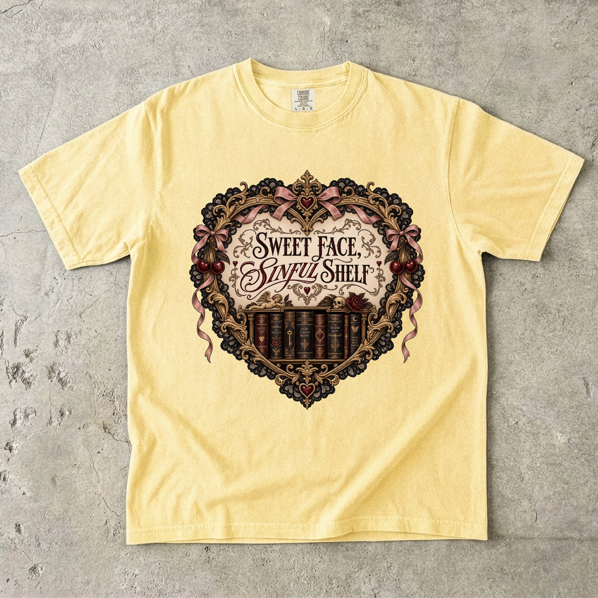 A pale yellow short-sleeve t-shirt laid flat on a textured gray concrete surface. The front of the shirt features a large, ornate gothic-style heart graphic with the text 'Sweet Face, Sinful Shelf' and an illustration of vintage book spines.