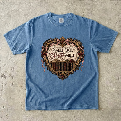 A light blue short-sleeve t-shirt laid flat on a textured concrete surface, featuring a large vintage-style graphic on the chest with a heart-shaped bookshelf design and the text 'Sweet Face and Sorry Shelf'.