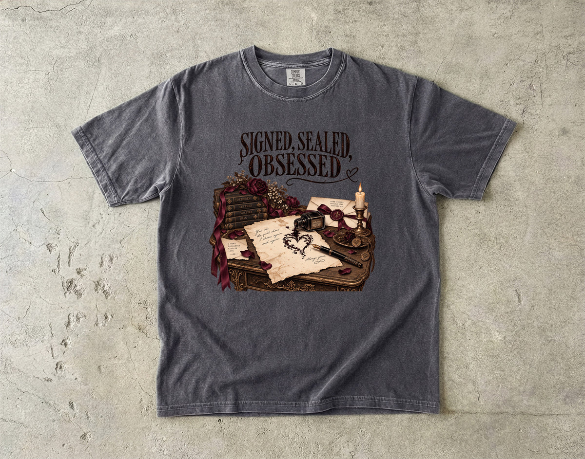 A dark gray heather t-shirt with a graphic print of a desk scene featuring a quill, inkwell, old letters, and roses, with the text 'SIGNED, SEALED, OBSESSED' above it. The t-shirt is laid flat on a textured concrete surface.