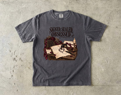 A dark gray heather t-shirt with a graphic print of a desk scene featuring a quill, inkwell, old letters, and roses, with the text 'SIGNED, SEALED, OBSESSED' above it. The t-shirt is laid flat on a textured concrete surface.