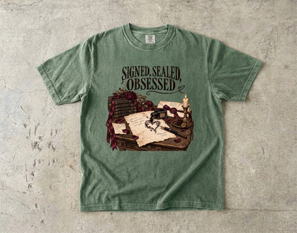 A sage green graphic t-shirt laid flat on a textured gray concrete surface. The shirt features a vintage-style illustration of a desk with parchment, ink, and a candle, with the text 'SIGNED, SEALED, OBSESSED' printed above the graphic.