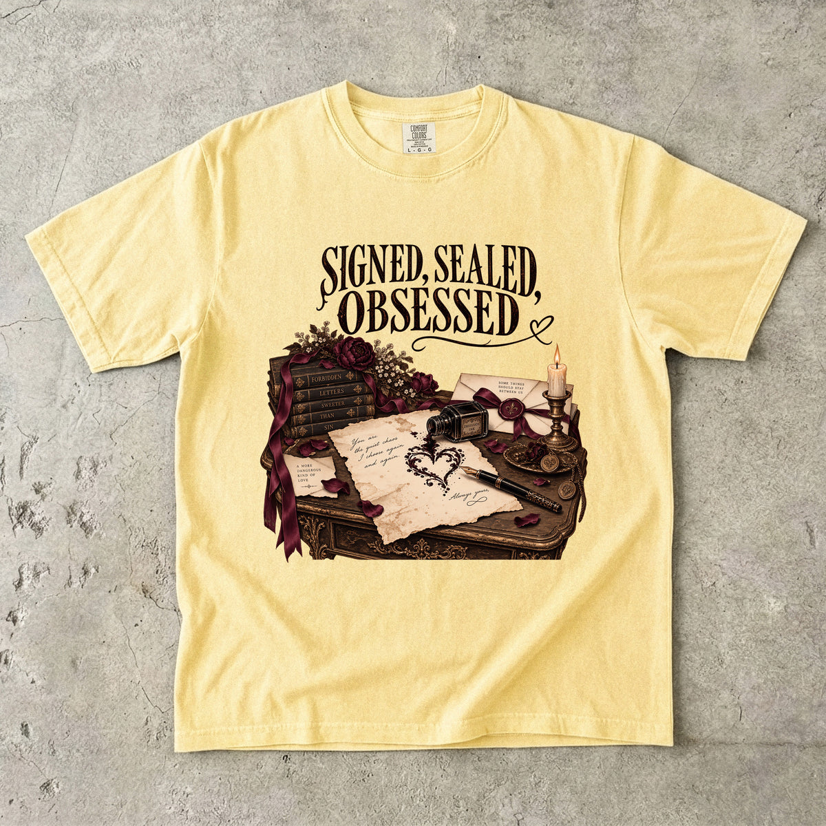 A pale yellow short-sleeve t-shirt laid flat on a textured gray concrete background. The shirt features a graphic print of a desk with old books, a quill, wax seals, and a candle, with the text 'SIGNED, SEALED, OBSESSED' printed above the illustration.