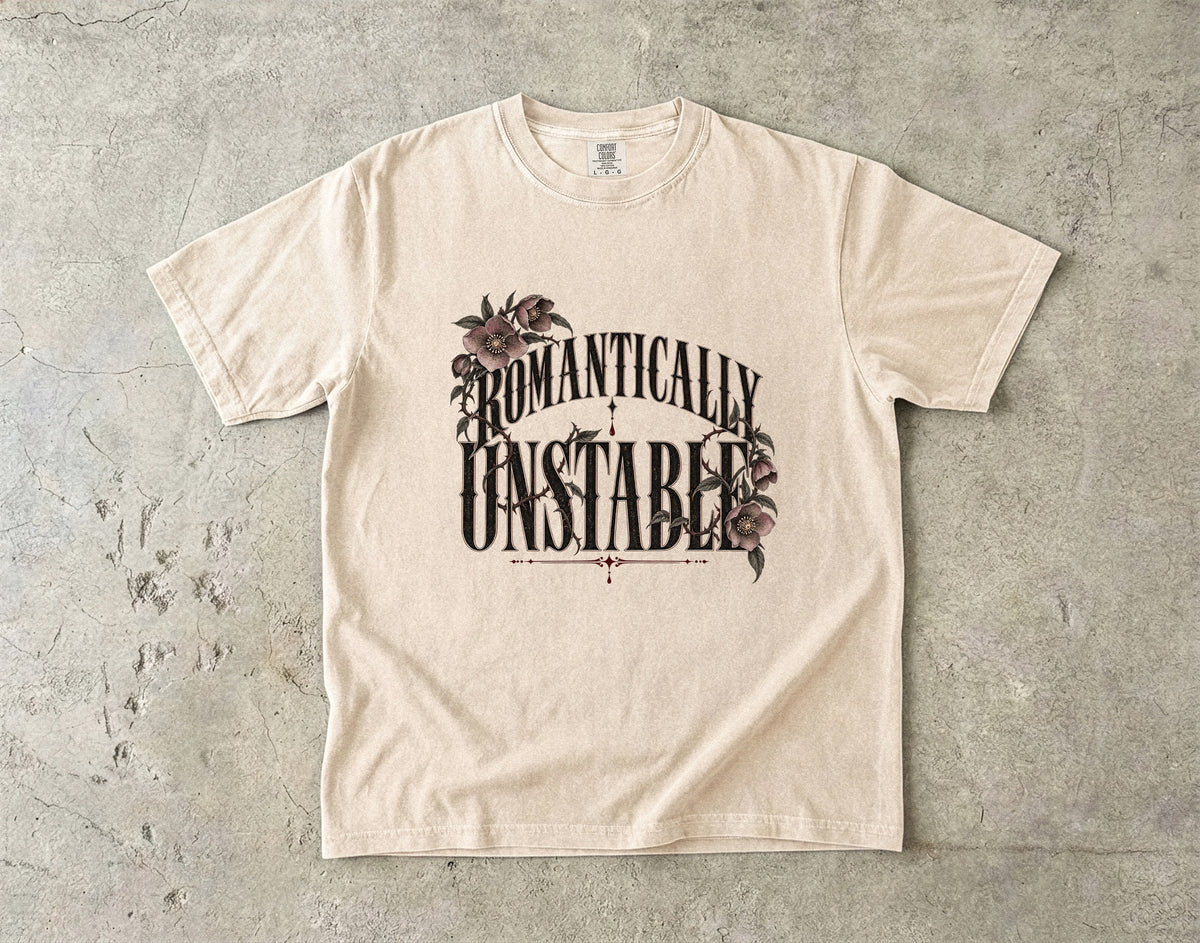 A cream-colored short-sleeve t-shirt with a graphic print reading 'ROMANTICALLY UNSTABLE' in black stylized lettering, decorated with dark brown floral illustrations, laid flat on a textured gray concrete surface.