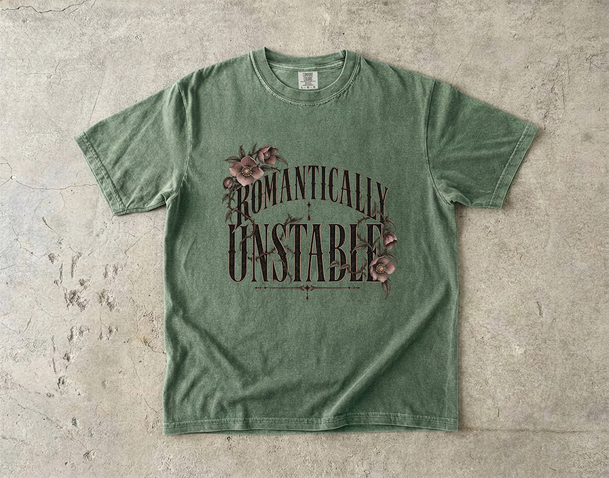 A heather green short-sleeve t-shirt with a vintage-style graphic reading 'ROMANTICALLY UNSTABLE' in dark brown lettering, decorated with pink roses, laid flat on a textured concrete surface.