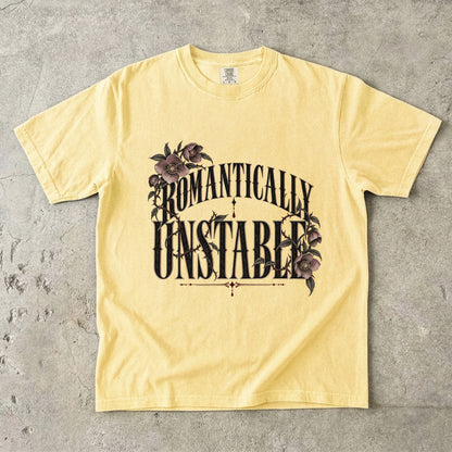 A pale yellow short-sleeve t-shirt with a graphic print reading 'ROMANTICALLY UNSTABLE' in black stylized lettering, decorated with dark purple flowers and green leaves, laid flat on a textured gray concrete surface.