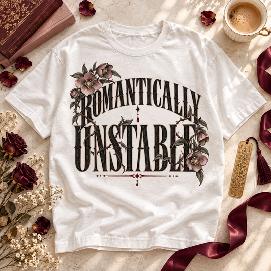 A white graphic t-shirt laid flat on a light marble surface, surrounded by styled props including dried roses, a book, a cup of coffee, a gold bookmark, and burgundy ribbon. The t-shirt features a gothic-style graphic reading 'ROMANTICALLY UNSTABLE' in black lettering with floral illustrations.