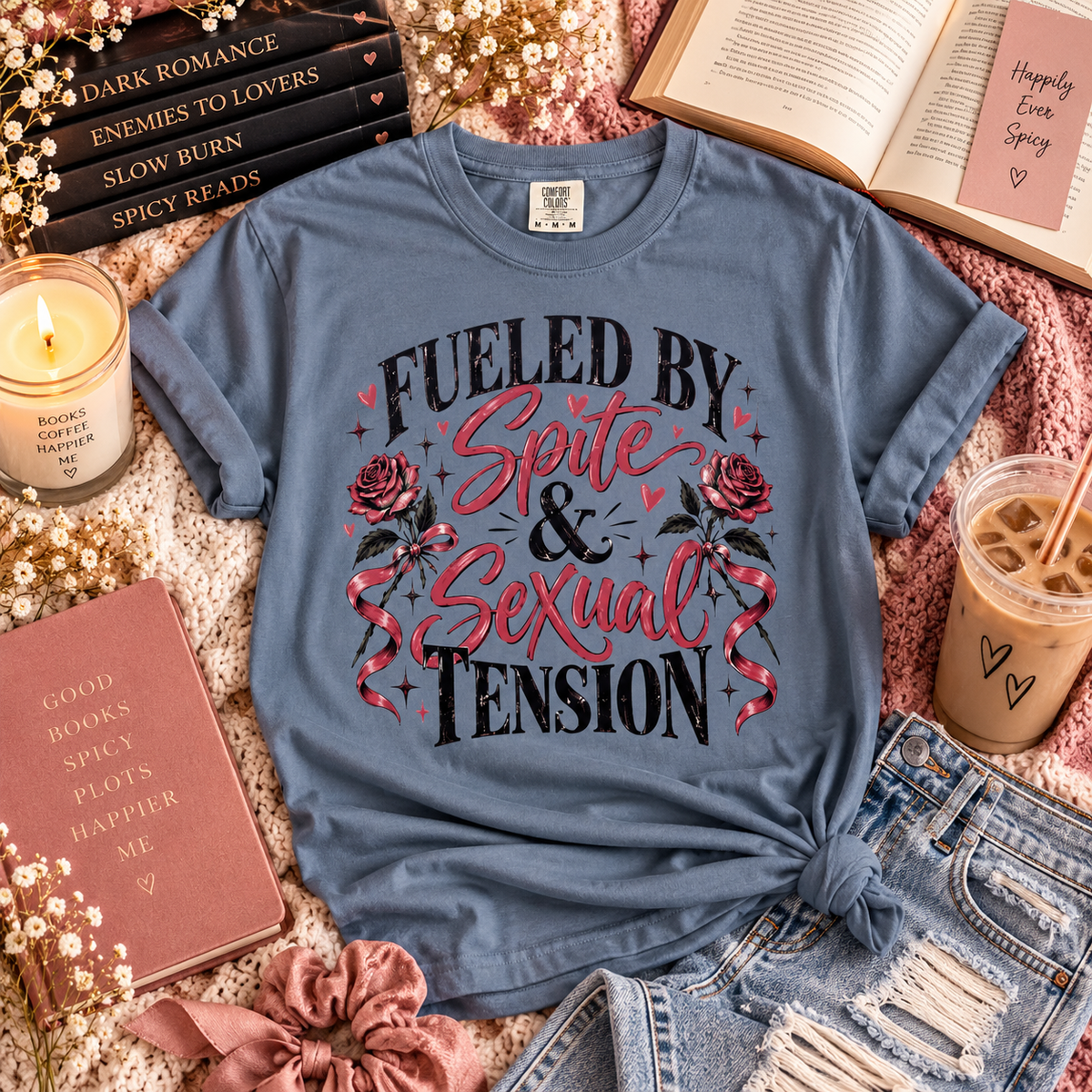 A slate blue graphic t-shirt with the text 'FUELED BY Spite & Sexual TENSION' and rose illustrations, styled in a cozy flat-lay arrangement with books, a candle, a drink, and denim shorts.