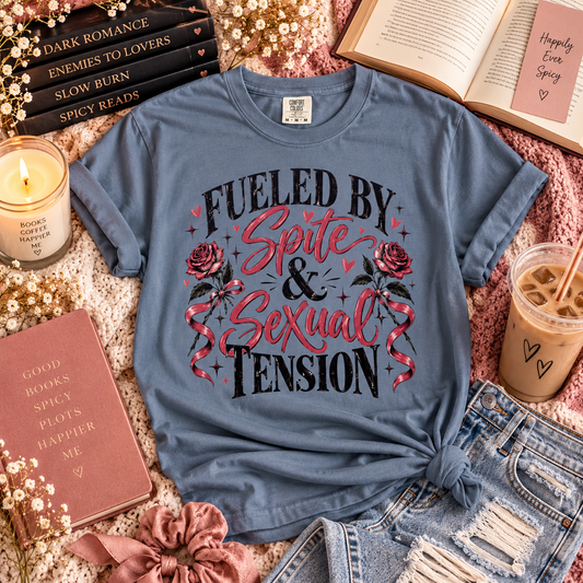 A slate blue graphic t-shirt with the text 'FUELED BY Spite & Sexual TENSION' and rose illustrations, styled in a cozy flat-lay arrangement with books, a candle, a drink, and denim shorts.