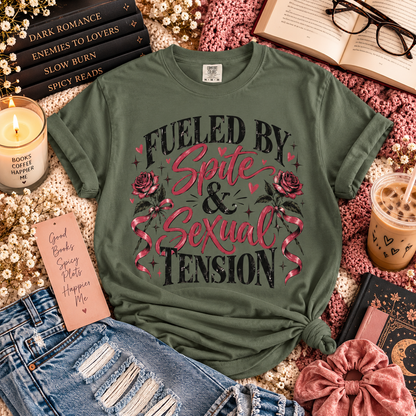 An olive green graphic t-shirt with the text 'FUELED BY Spite & Sexual Tension' and pink roses, styled in a cozy flat-lay arrangement with jeans, a candle, an iced coffee, and books.