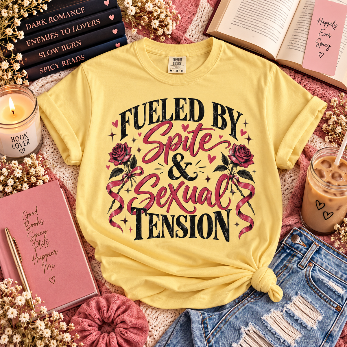 A yellow graphic T-shirt laid flat in a styled flat-lay arrangement with books, a candle, a notebook, an iced coffee, and denim shorts. The shirt reads 'FUELED BY Spite & Sexual Tension' with pink roses and black text.