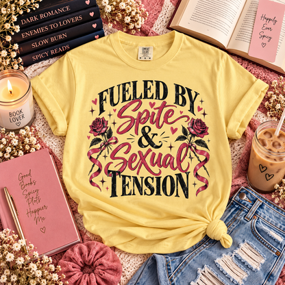 A yellow graphic T-shirt laid flat in a styled flat-lay arrangement with books, a candle, a notebook, an iced coffee, and denim shorts. The shirt reads 'FUELED BY Spite & Sexual Tension' with pink roses and black text.