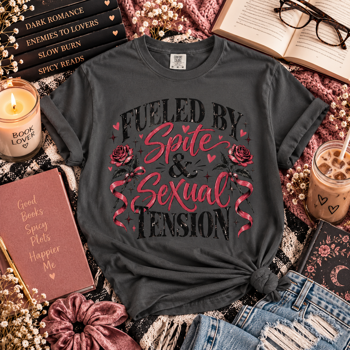 A dark charcoal gray graphic t-shirt with a romantic, cozy-themed flat-lay arrangement. The t-shirt features the text 'FUELED BY Spite & Sexual TENSION' in black and pink lettering, decorated with pink roses and hearts.