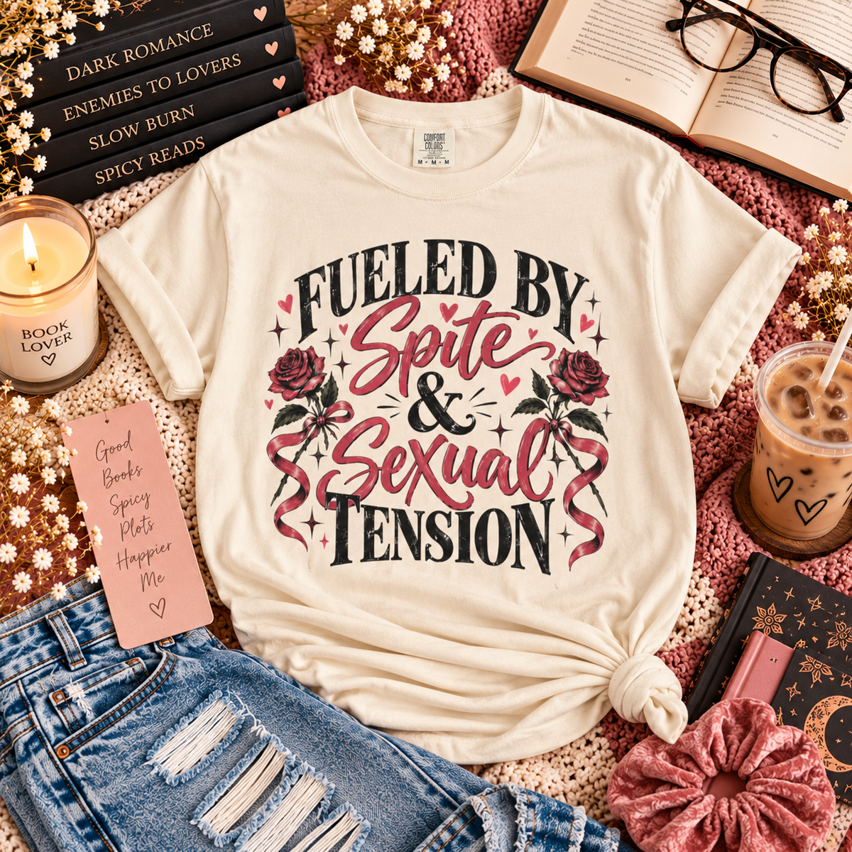 An off-white graphic t-shirt with the text 'FUELED BY Spite & Sexual Tension' and rose illustrations, styled in a cozy flat-lay arrangement with jeans, a candle, an iced coffee, and books.