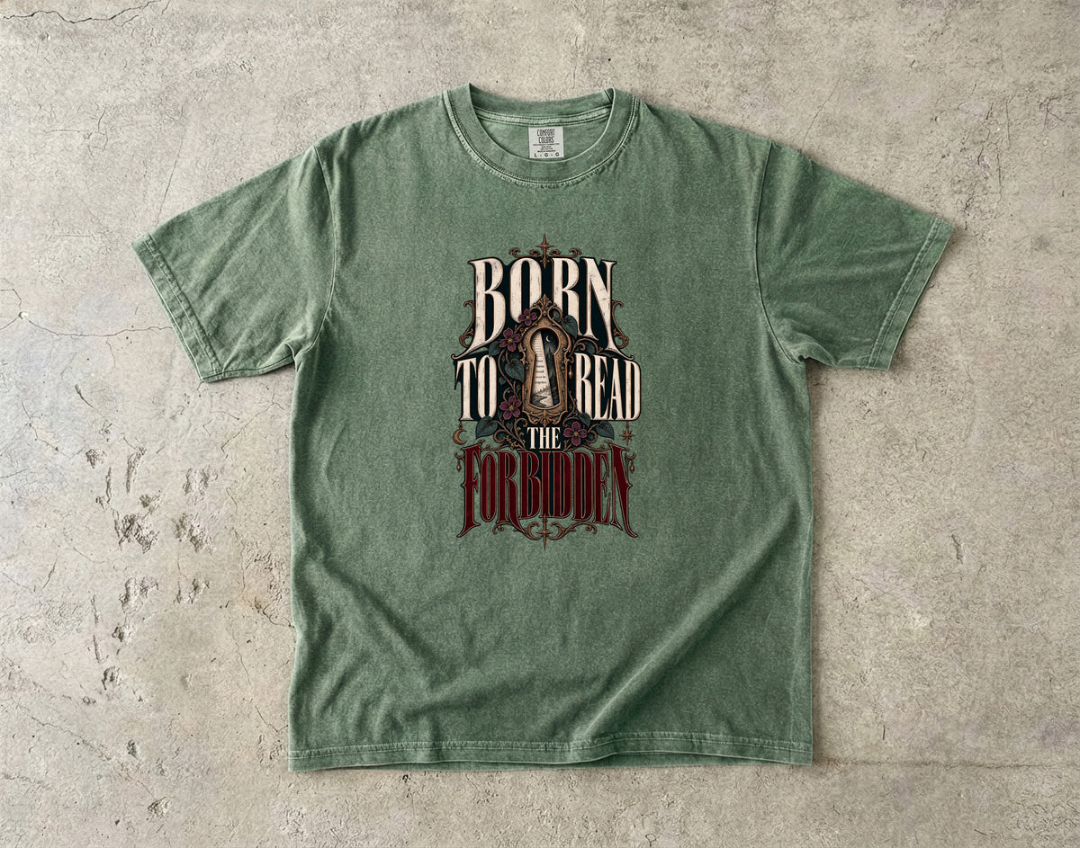 A green short-sleeve graphic t-shirt laid flat on a textured concrete surface. The shirt features a distressed graphic on the front with the words 'BORN TO READ THE FORGOTTEN' in stylized lettering.