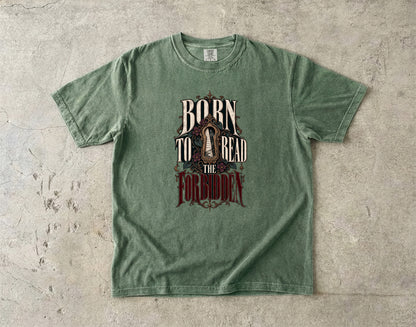 A green short-sleeve graphic t-shirt laid flat on a textured concrete surface. The shirt features a distressed graphic on the front with the words 'BORN TO READ THE FORGOTTEN' in stylized lettering.