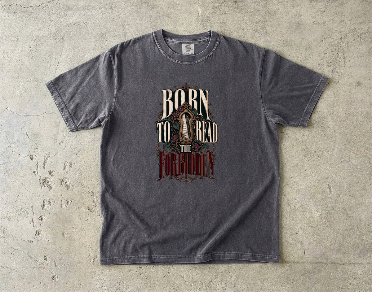 A dark gray crewneck t-shirt with a distressed graphic reading 'BORN TO READ THE FORGOTTEN' in cream and red lettering, laid flat on a textured concrete surface.