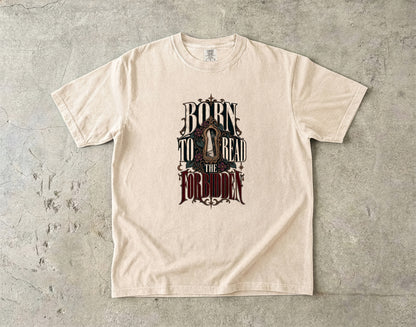 A cream-colored short-sleeve t-shirt with a gothic-style graphic reading 'BORN TO READ THE FORBIDDEN' in dark lettering, laid flat on a textured gray concrete surface.