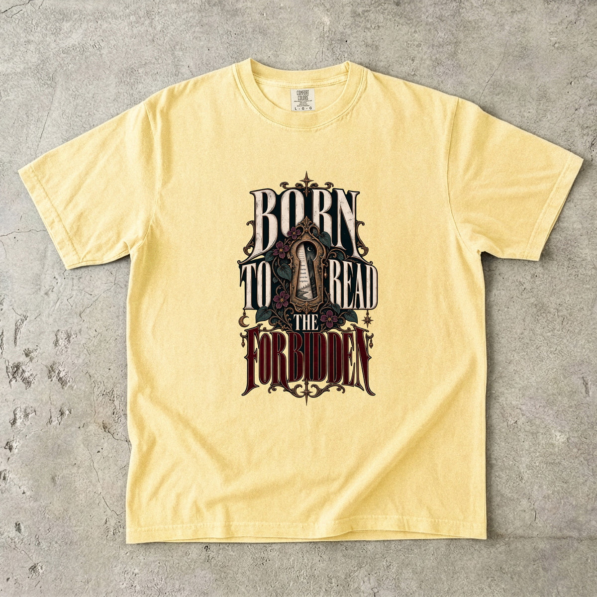 A pale yellow short-sleeve t-shirt with a gothic-style graphic reading 'BORN TO READ THE FORBIDDEN' in dark lettering, laid flat on a textured gray concrete surface.