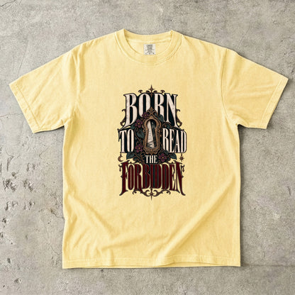 A pale yellow short-sleeve t-shirt with a gothic-style graphic reading 'BORN TO READ THE FORBIDDEN' in dark lettering, laid flat on a textured gray concrete surface.