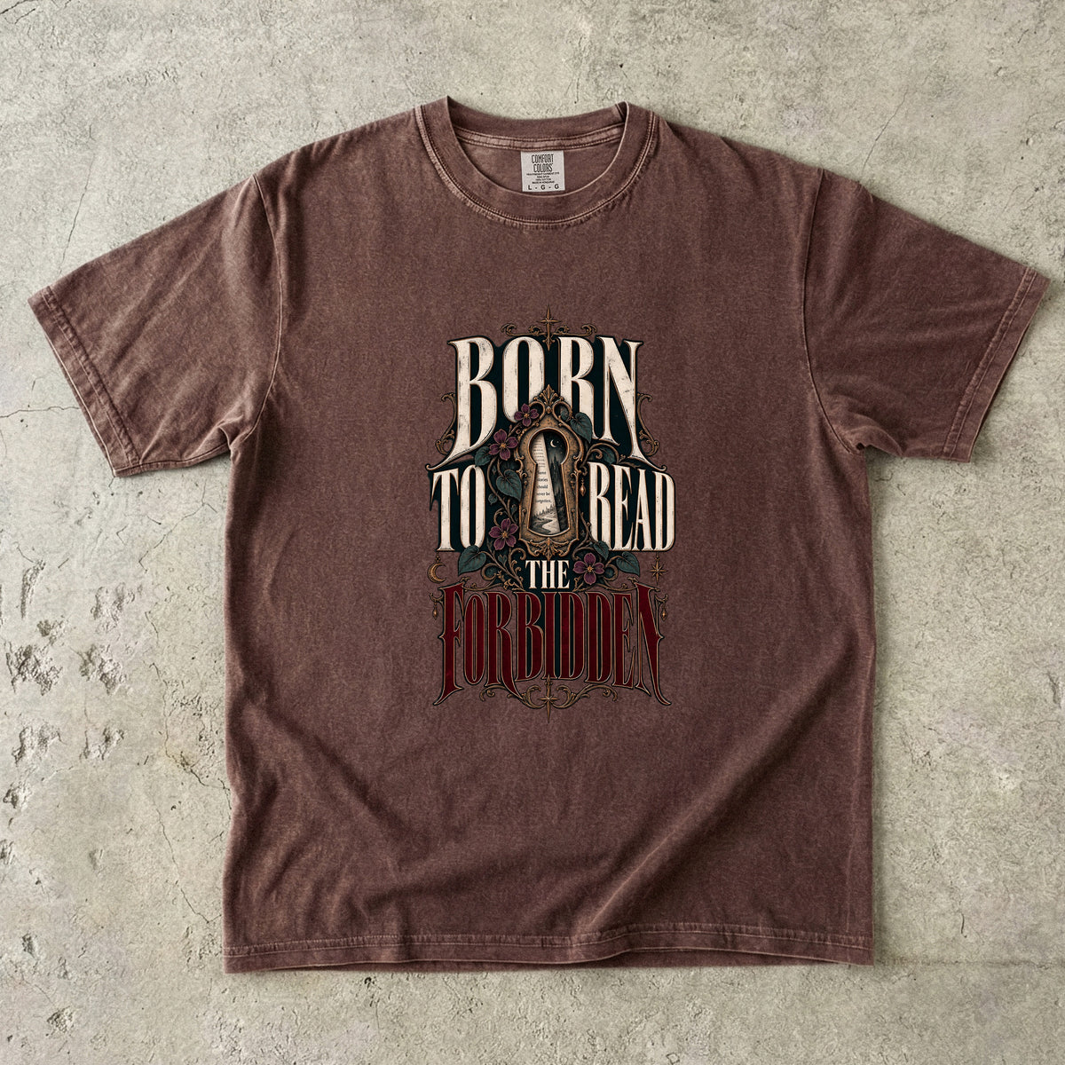 A brown short-sleeve graphic t-shirt laid flat on a textured gray concrete surface. The shirt features a distressed graphic on the front with the text 'BORN TO READ THE FORBIDDEN' in stylized cream and red lettering.
