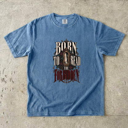 A light blue short-sleeve graphic t-shirt laid flat on a textured concrete surface. The graphic features the text 'BORN TO READ THE FORBIDDEN' in a stylized, vintage font with a dark, ornate illustration in the center.