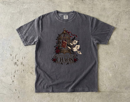 A dark gray crewneck t-shirt with a graphic print of a birdcage, red roses, flying parchment scrolls, and the text 'BURN THE CHAOS' laid flat on a textured concrete surface.