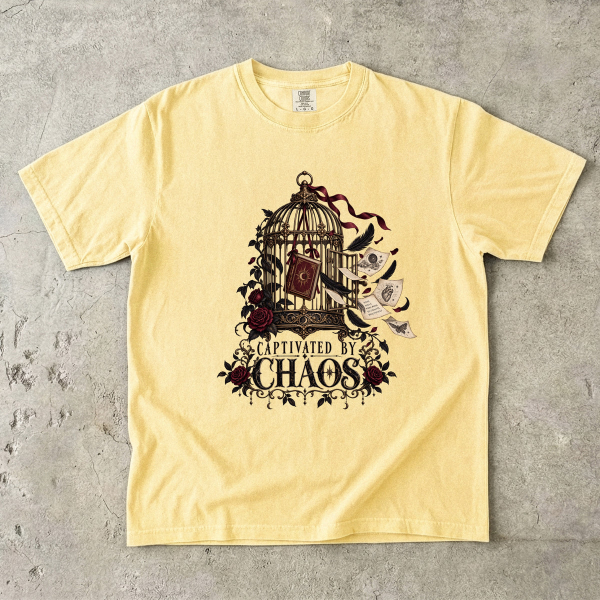 A pale yellow short-sleeve t-shirt with a gothic-style graphic print of a birdcage, roses, a book, and a ghost, with the text 'CAPTIVATED IN CHAOS', laid flat on a textured gray concrete surface.