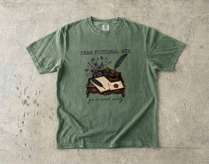 A sage green graphic t-shirt laid flat on a textured concrete surface. The shirt features a vintage-style illustration of a desk with an open book, inkwell, and quill, with the text 'DEAR FICTIONAL MEN, you are reality' printed on the front.